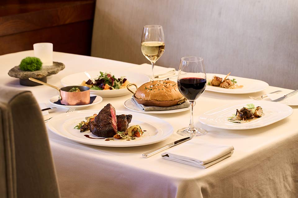 a dinner table with a fine dining steak dinner, salads, bread, and glasses of red and white wine